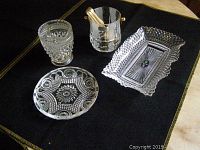 Four assorted glass serving dishes displayed on black surface: rectangular tray, round plate, faceted glass container, small metal bucket with wooden tongs.