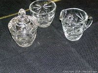Clear glass creamer, covered sugar bowl, and small bowl all featuring star cut patterns photographed on a dark surface.