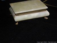 Front-side angle showing the keepsake box with light cream colored stone and metal claw feet on a black background.