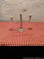 Photo of a silver-colored metal three-arm candle holder with detailed base on a red and white checkered surface against a textured wall.