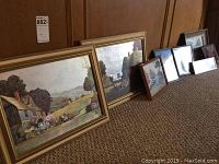 Seven framed landscape paintings with a variety of countryside scenes plus one empty frame all placed on carpeted floor against wood panel wall.