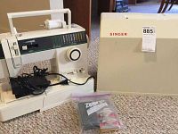 White Singer portable sewing machine next to beige carrying case and plastic bag with accessories on carpeted floor
