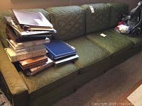 Green fabric sofa with three seat cushions and three back cushions, stacked with books and other items on the left side.