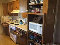 Wide view showing kitchen countertop with stove, microwave, open drawers containing kitchen utensils and various cookware on stove and counter