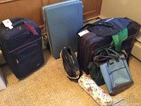 Five pieces of luggage and various travel bags arranged on floor including hard and soft cases and bags of different sizes and colors.