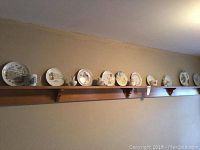 Wide view of the collection of 19 plates and dishes mounted on a wooden wall shelf, showing assorted decorative and commemorative plates in round shapes.
