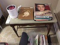 Wooden piano bench with a cushioned beige seat and a stack of sheet music books on top, including one with Elvis Presley on the cover and another with Betty Clive.