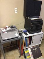 Photo of black GE stereo system components stacked with black speakers on top, next to white cabinet holding vinyl records and a Brother typewriter on a small table.