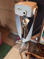 Full view of vintage grey and beige standing belt massager with control knob, fabric belt, on broad base, placed on carpeted floor.