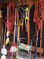 Photo showing various handled cleaning tools including brooms, mop, dustpan, and a green metal step ladder with two steps, along with orange extension cords hanging on the wall.