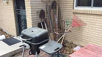 Wide view showing the UniFlame charcoal grill, assorted yard tools leaning against the wall, leaf bag cart, and outdoor umbrella stand.