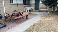 Image of reddish-brown wooden picnic table with two matching benches on brick patio against house wall.