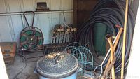 Photo showing garden hoses coiled on reels, wooden lawn croquet mallets, metal trash can with lid, and some garden tools against shed wall.