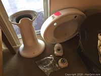 Old white ceramic pedestal sink bowl, pedestal stand, and drainage accessories on floor next to glass door. Sink bowl has rounded edges and mounting holes. Pedestal is cylindrical with flared base.