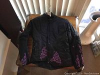 Full front view of black Joe Rocket motorcycle jacket with pink embroidery and zipper closure, displayed on wooden chair.