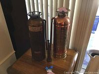 Photo of two vintage fire extinguishers placed on a wooden table near vertical blinds, showing overall appearance and size.