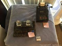 Vintage black mechanical calculator and a desk set with glass ink bottles on a cloth-covered table.