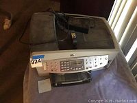 Front view of HP OfficeJet 7210 all-in-one printer on a gray surface showing keypad and LCD screen, power cord and ink cartridge beside it.