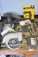 Shows Craftsman circular saw, electric sander, clamps, extension cord, and drill chucks arranged on a wooden work surface.