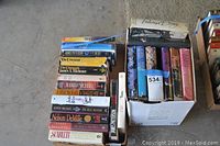 Two boxes of mystery and adventure books from various authors including James Patterson, Judith Michael, Judith Krantz, Jackie Collins, and Nelson DeMille. Books are both paperback and hardcover with some dust jackets.