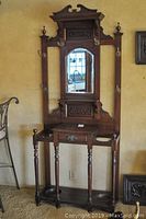 Full view of the antique wooden hall tree showing mirror, shelf, drawer, hooks, and turned legs.