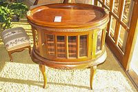 Front view of antique wooden tray table with removable tray top and glass-encased cabinet below. Oval shape with carved decorative edge and curved legs.