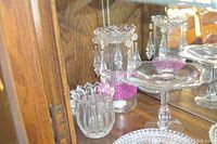 Glassware displayed in wooden cabinet with mirrored back. Includes pedestal stemmed bowl, decorative crystal prism container, clear glass bowls, and plates visible.