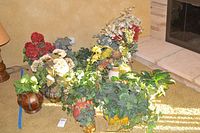 14 faux flower and foliage plants arranged near a corner fireplace with light from window illuminating them.