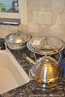 Two aluminum chafing bowls with lids and a polished stainless steel teapot placed on granite countertop near kitchen sink.