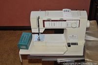 Off-white Husqvarna sewing machine with stitch selector and dial on the front, placed on a beige surface with a turquoise box labeled 'Rotary Attachments' next to it.