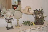 Photo showing two lamps (one large floral glass design and one smaller brass with frosted glass shade), straw hat on metal stand, decorative birdcage with artificial flowers, and some artificial greenery on surface.