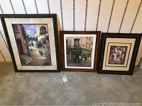 Three framed cityscape art prints leaning against a wall on carpet, showing varying sizes and frame styles