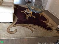 Photo showing large rug under glass table and beige couch, displaying swirled floral patterned edges with beige background.