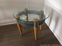 Oval glass top side table with wood legs and gray metal accents, shown from an angle.