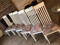 Six white wooden dining chairs aligned side by side showing vertical slatted backs and patterned padded seats.