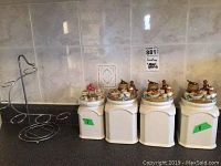 Set of four white ceramic canisters with decorative lids placed on a countertop showing three tall canisters of 8 inches and one smaller canister 7 inches tall. All lids have kitchen-themed sculptural details with miniature kitchen items in various colors.