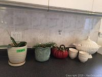 White ceramic soup tureen with ladle, six matching bowls, tomato-shaped pitcher, green and beige planters all on kitchen counter, showing minor chips on planters and pitcher