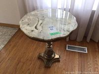 Full view of the round marble topped table with gold toned pedestal base placed on wooden floor near window.
