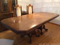 Full view of large oak dining table with polished octagonal top and visible wood grain, surrounded by dining chairs and cabinet in background.