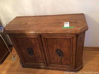 Front view of oak wood cabinet, showing two closed doors with brass ring pull handles and beveled top surface.