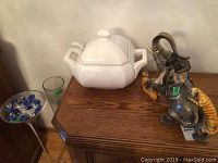 Wide view showing white ceramic soup tureen with ladle, elephant figurine with two tigers and native rider, both on wooden surface with glass display of stones to the side