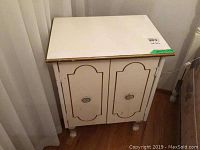 Front and angled views of white night table with gold edging, showing two paneled doors with metal knobs and signs of wear on surface and edges.