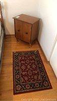 Photo of wooden night table cabinet with round knobs beside a 2x3 feet burgundy floral area rug
