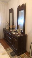 Photo of wood dresser with two attached tall mirrors, decorative carvings on drawer fronts visible.