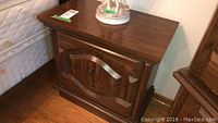 First night table showing carved wooden front panel, two metal knob handles, minor surface wear, placed beside a bed.