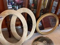 Photo showing two cream-colored oval frames with raised floral carvings and one empty gold oval frame with mirror leaning against a wooden cabinet.