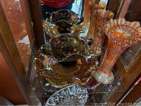 Photo showing two orange carnival glass vases and a two-handled carnival glass platter with floral and grape designs and a cut glass bowl underneath.