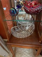 Red cut glass bowl, clear cut glass bowl, and rainbow snail figurine displayed inside a wooden and glass cabinet.