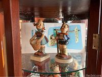 Two Hummel children figurines on a glass shelf, one girl with a basket, one boy with a basket.