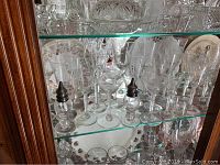 Glass cabinet shelf showing a group of multiple cut glass stemware with etched patterns including wine and martini glasses. Two small glass salt and pepper shakers also visible.